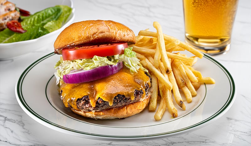 Cheeseburger with lettuce, tomato, onion, fries, and a glass of beer on a plate, next to a salad—perfect for enjoying in the comfort of our stylish rooms.