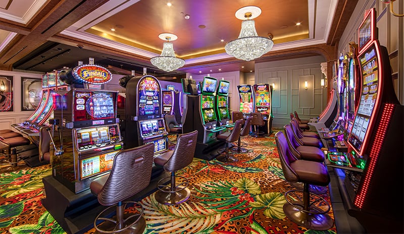 A lively casino room with colorful slot machines, brown chairs, chandeliers, and a vibrant patterned carpet creates one of the most inviting rooms for entertainment and excitement.